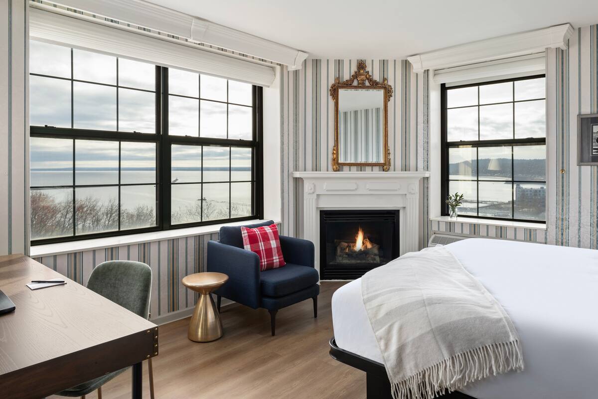 A hotel room with a large bed, a fireplace, a seating area with a chair and a small table, and large windows offering a view of the outdoors.
