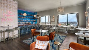 A modern bar area with a blue wall, a variety of bottles, and a neon sign. There are multiple seating areas with chairs and tables, and a large window offering a view of the city. - Landmark Inn (Marquette)