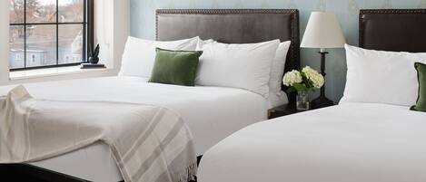 A hotel room with two beds, a window with a view, and a nightstand with a lamp and flowers.