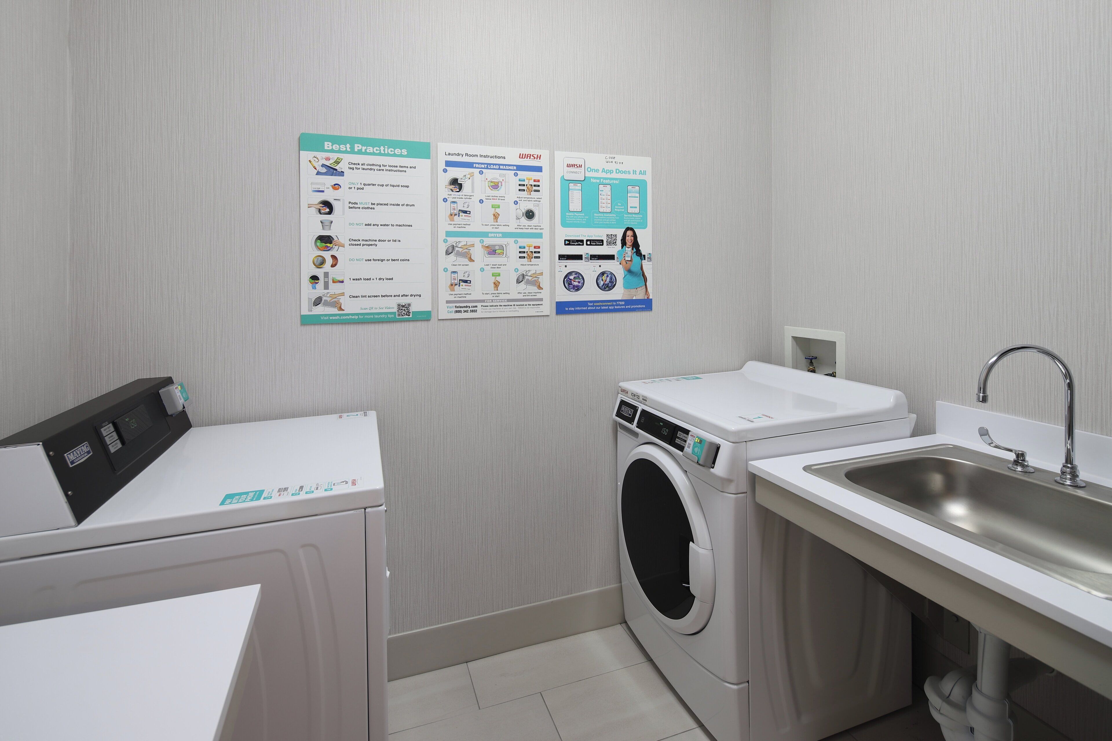 A laundry room with a front-loading washer, a dryer, and a sink. There are instructional posters on the wall.