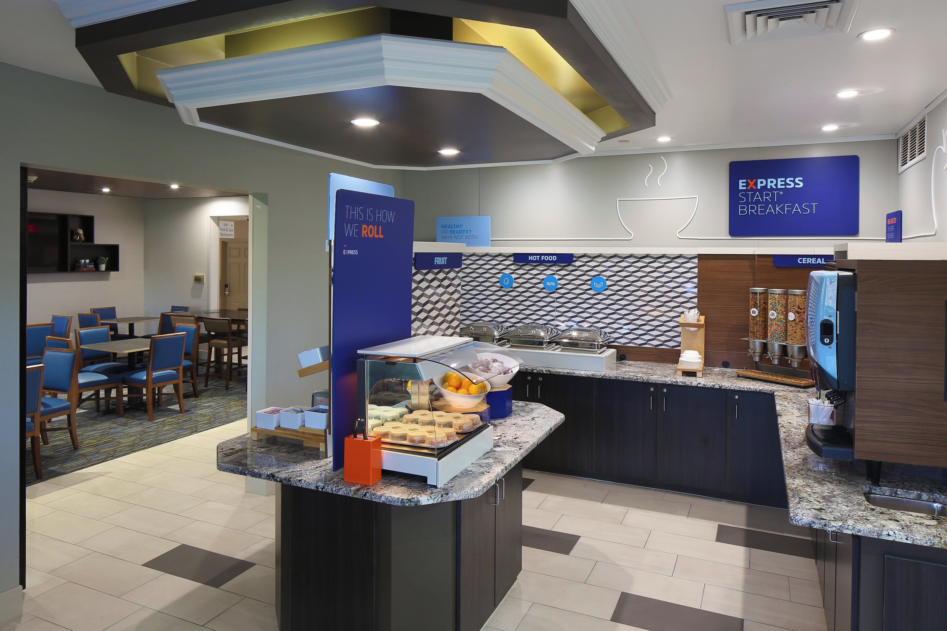 A hotel breakfast area with a self-service counter, seating area, and signage indicating amenities like a pool and spa.