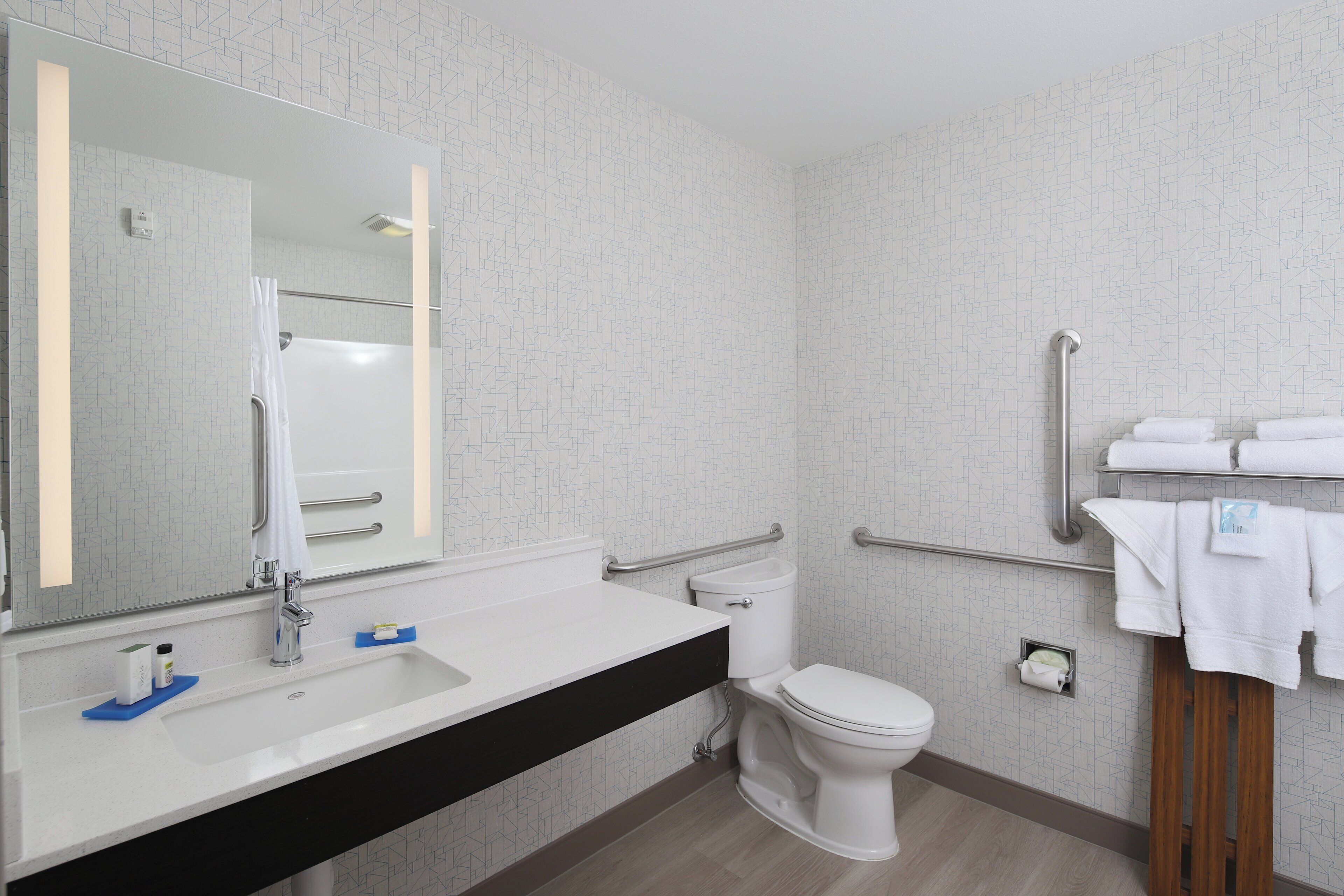 Een moderne badkamer met een wit wastafelmeubel, spiegel en toilet. Er zijn handgrepen en een handdoekhouder.