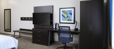 A hotel room with a bed, a desk with a flat-screen TV, a chair, a wardrobe, and a framed picture on the wall.