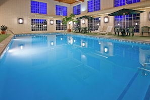 An indoor swimming pool with clear blue water, surrounded by lounge chairs and umbrellas, and a tiled wall with windows.