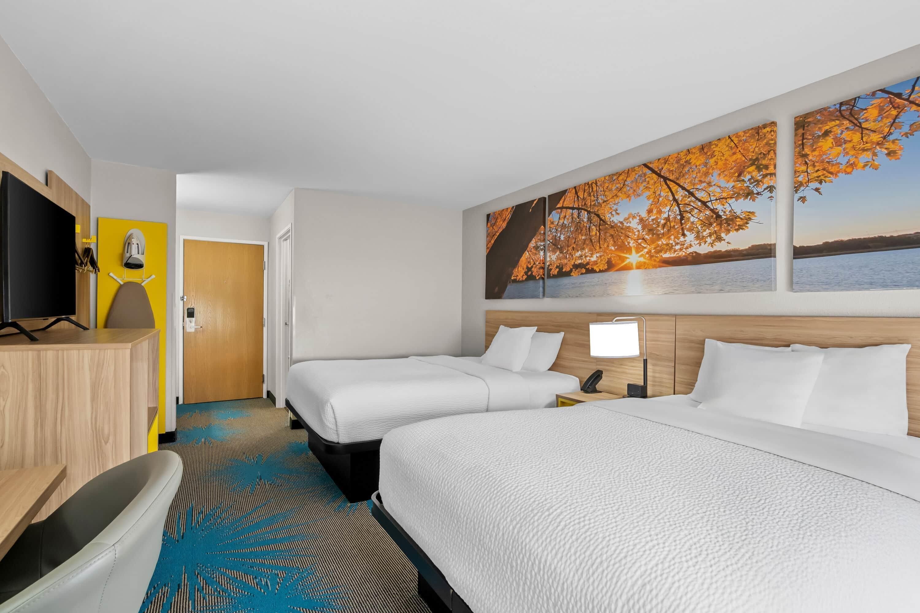 A hotel room with two beds, a desk, a TV, and a large window with a view of a lake and autumn leaves.