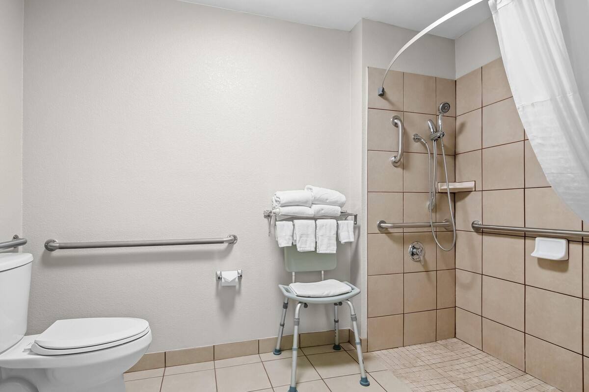 A bathroom with a toilet, a shower with grab bars, a wheelchair-accessible bench, and a towel rack.