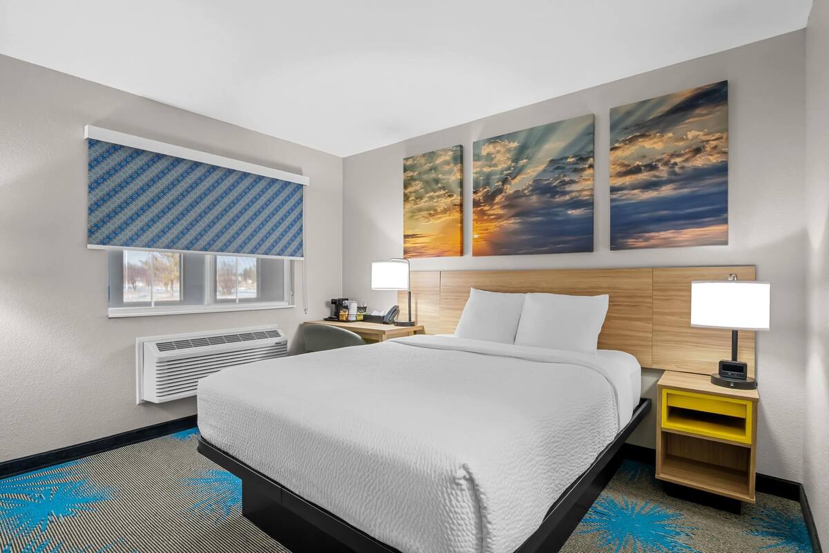 A hotel room with a large bed, a desk with a lamp, a window with blinds, and artwork on the wall.