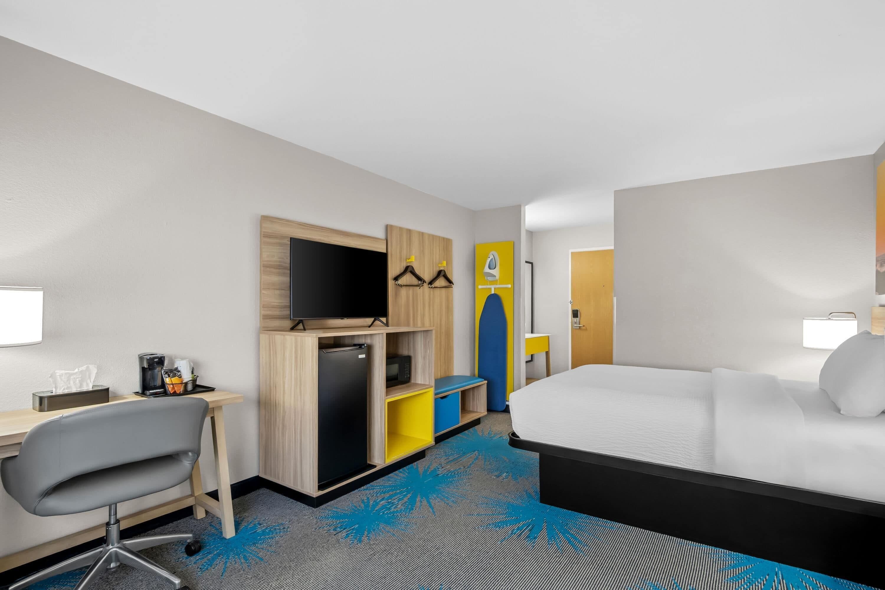 A hotel room with a bed, a desk with a chair, a television, a mini-fridge, and a wardrobe.