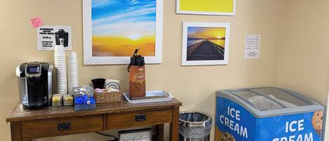 Un coin café équipé d’une machine à café, de tasses et d’un panneau indiquant où acheter du café. On y trouve également des tableaux encadrés au mur, une poubelle et un réfrigérateur pour les glaces.