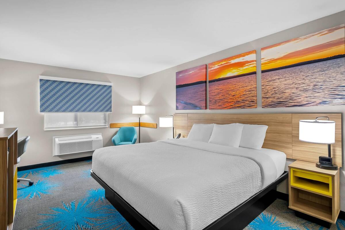 A hotel room with a large bed, a desk, a chair, a nightstand, and a wall with three sunset paintings.