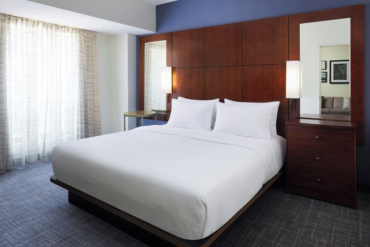 A hotel room with a large bed, a wooden headboard, a bedside table, a mirror, and a view of the outside through the window.