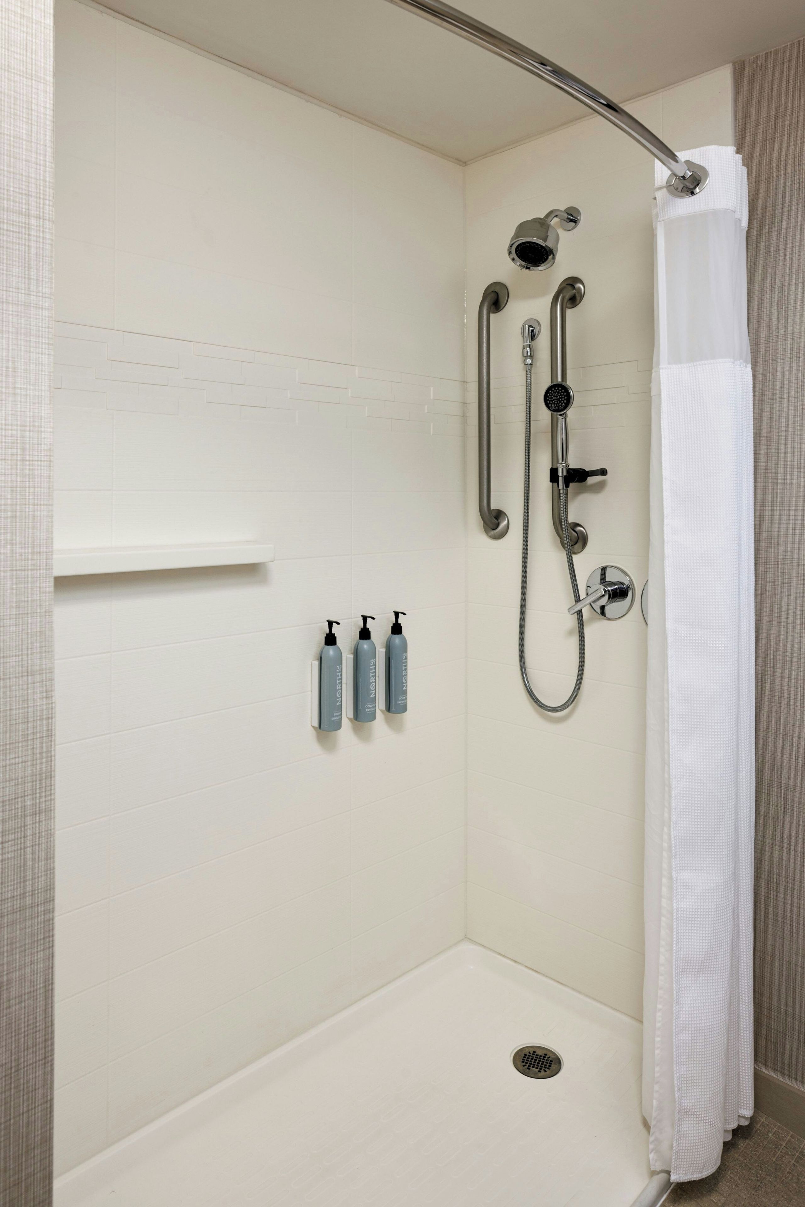 A walk-in shower with a showerhead, handheld shower, and a handheld shower attachment. There are three bottles of shampoo and conditioner on the wall.