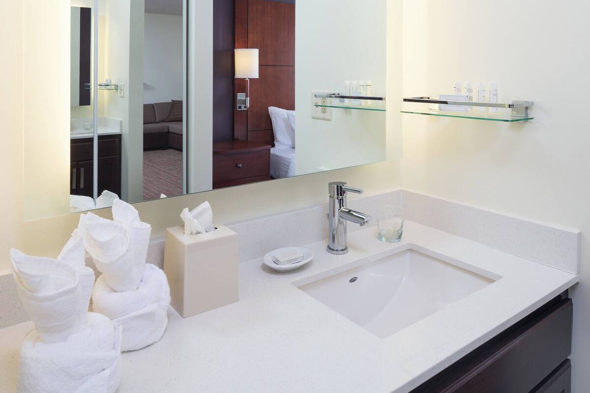 Une salle de bain d’hôtel avec un grand miroir, un lavabo blanc, une étagère en verre où sont posés les articles de toilette et des serviettes soigneusement pliées.