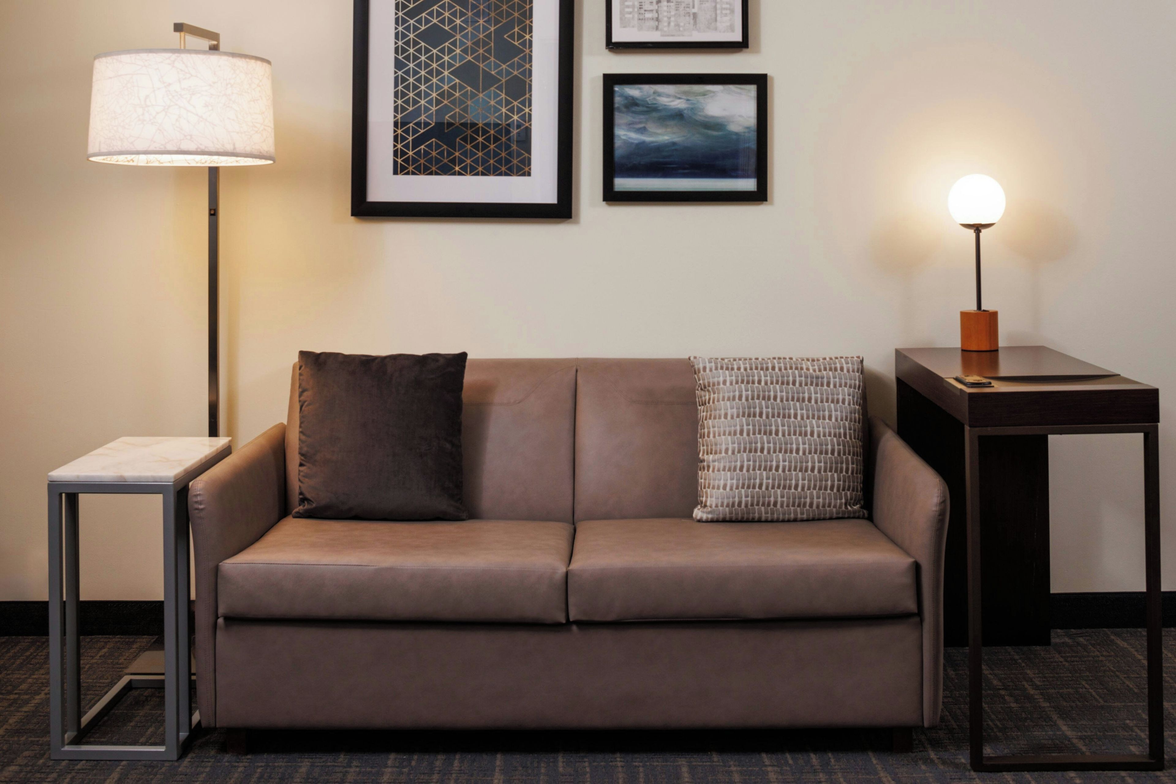 A room with a brown sofa, two pillows, a small side table, a lamp, and two framed pictures on the wall.