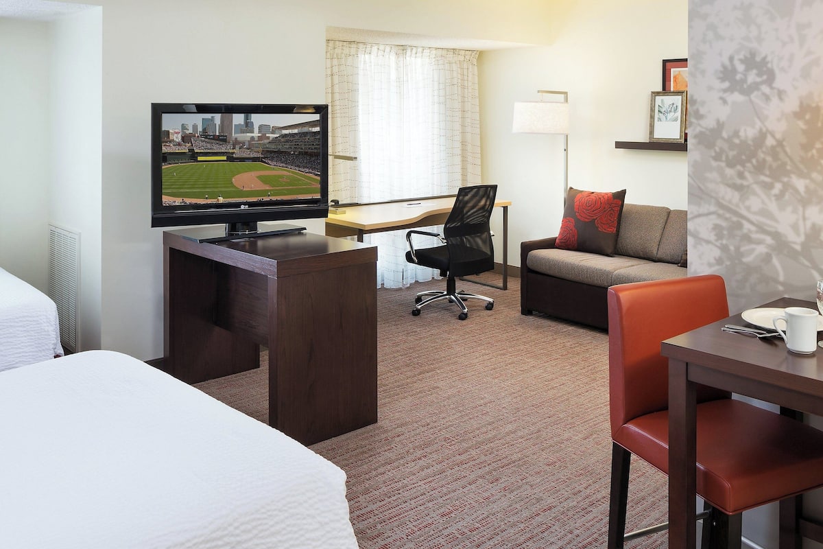 Une chambre d’hôtel avec un lit, un bureau sur lequel est posée une télévision diffusant un match de baseball, un canapé et une table à manger avec une tasse et une assiette.
