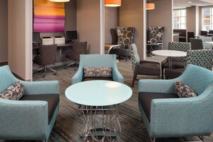 Un moderno vestíbulo de hotel con sillones azul verdosos, una mesa de centro de vidrio y una zona de recepción con computadora e impresora.