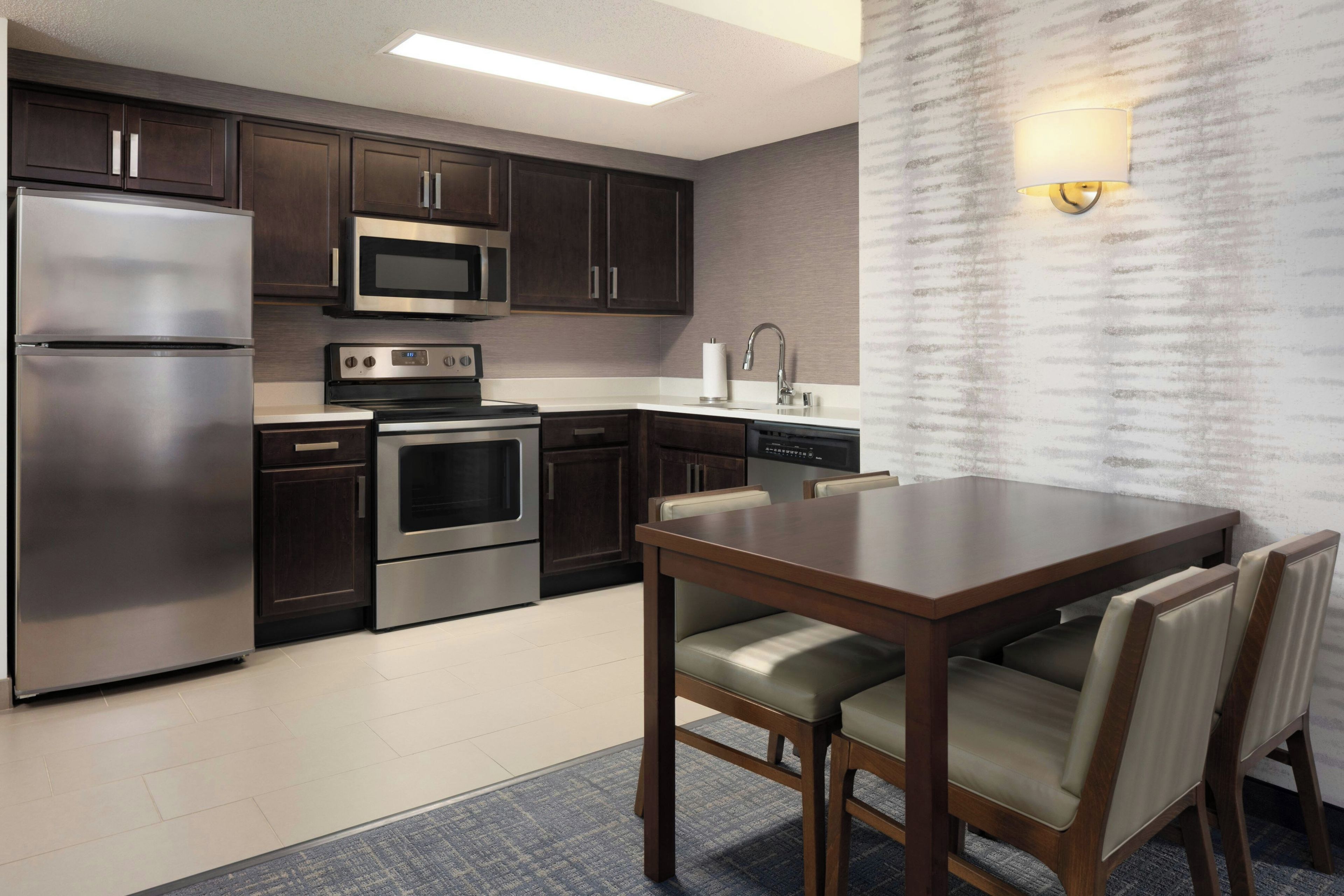 Una cocina moderna con electrodomésticos de acero inoxidable, armarios de madera oscura, microondas integrado y un comedor con mesa y sillas de madera.