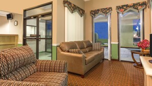 A hotel room with a brown patterned sofa, a matching armchair, a wooden coffee table, and a view of the outdoors through a glass door.