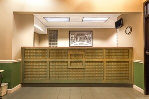 A reception area with a tiled floor, a reception desk with a framed picture, a clock, and a television mounted on the wall. - Super 8 by Wyndham Madison/Ridgeland Area (Madison)