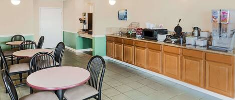 A breakfast area with tables and chairs, a counter with coffee machines, and a refrigerator with beverages.