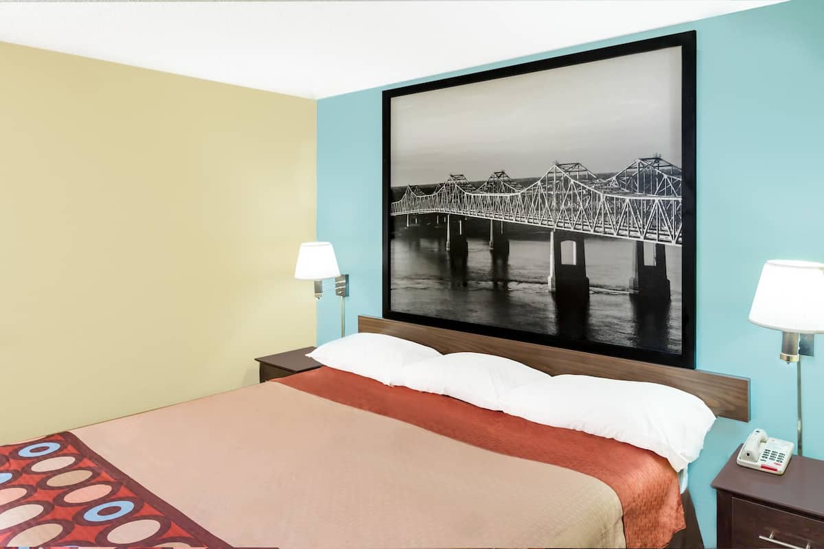Une chambre d’hôtel avec un lit, une table de chevet, un téléphone et un tableau représentant un pont, accroché au mur.