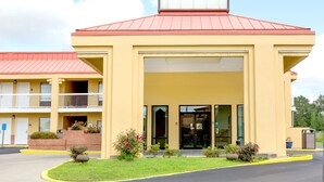 A two-story motel with a red roof and yellow walls, featuring a central entrance with glass doors and a small garden area with flowers.