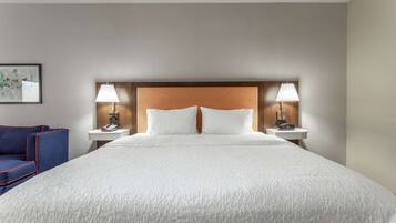 A hotel room with a large bed, two bedside tables, a nightstand, a blue chair, and a painting on the wall.
