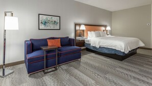 A hotel room with a bed, a sofa, a nightstand, a lamp, and a painting on the wall. - Hampton Inn Jefferson City at Capital Mall (Jefferson City)