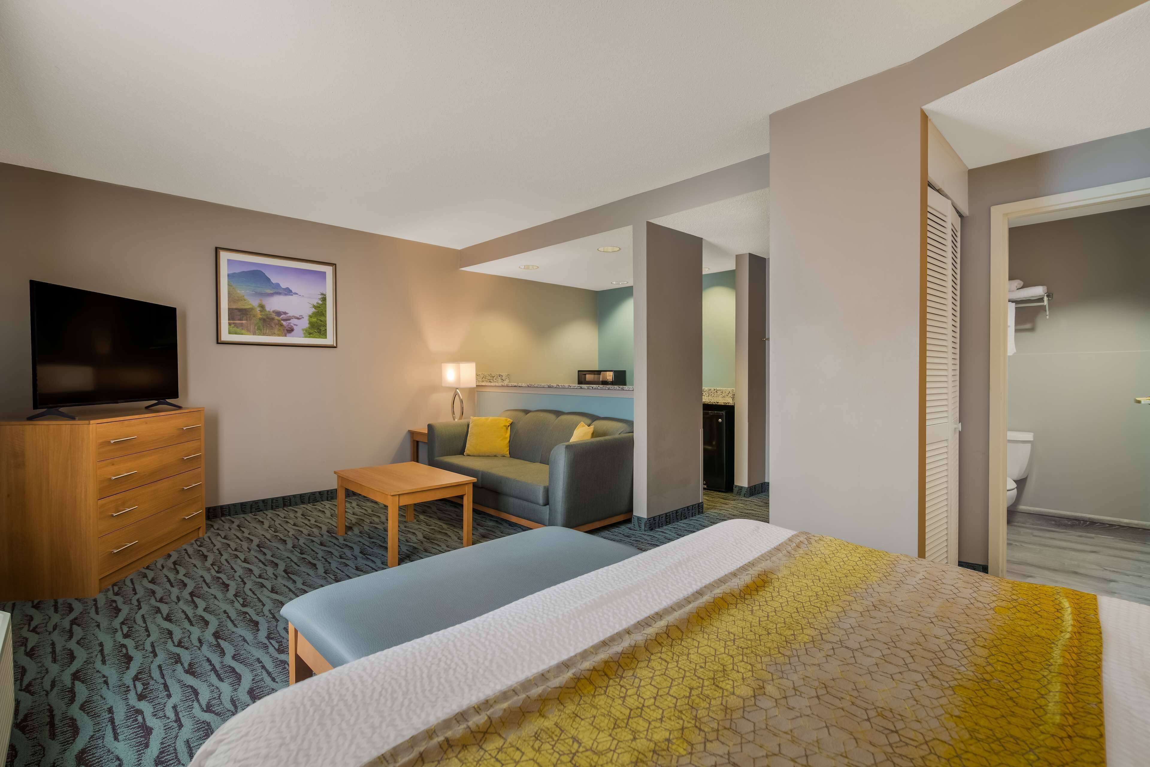 A hotel room with a bed, a TV, a sofa, a small table, and a bathroom visible through an open door.