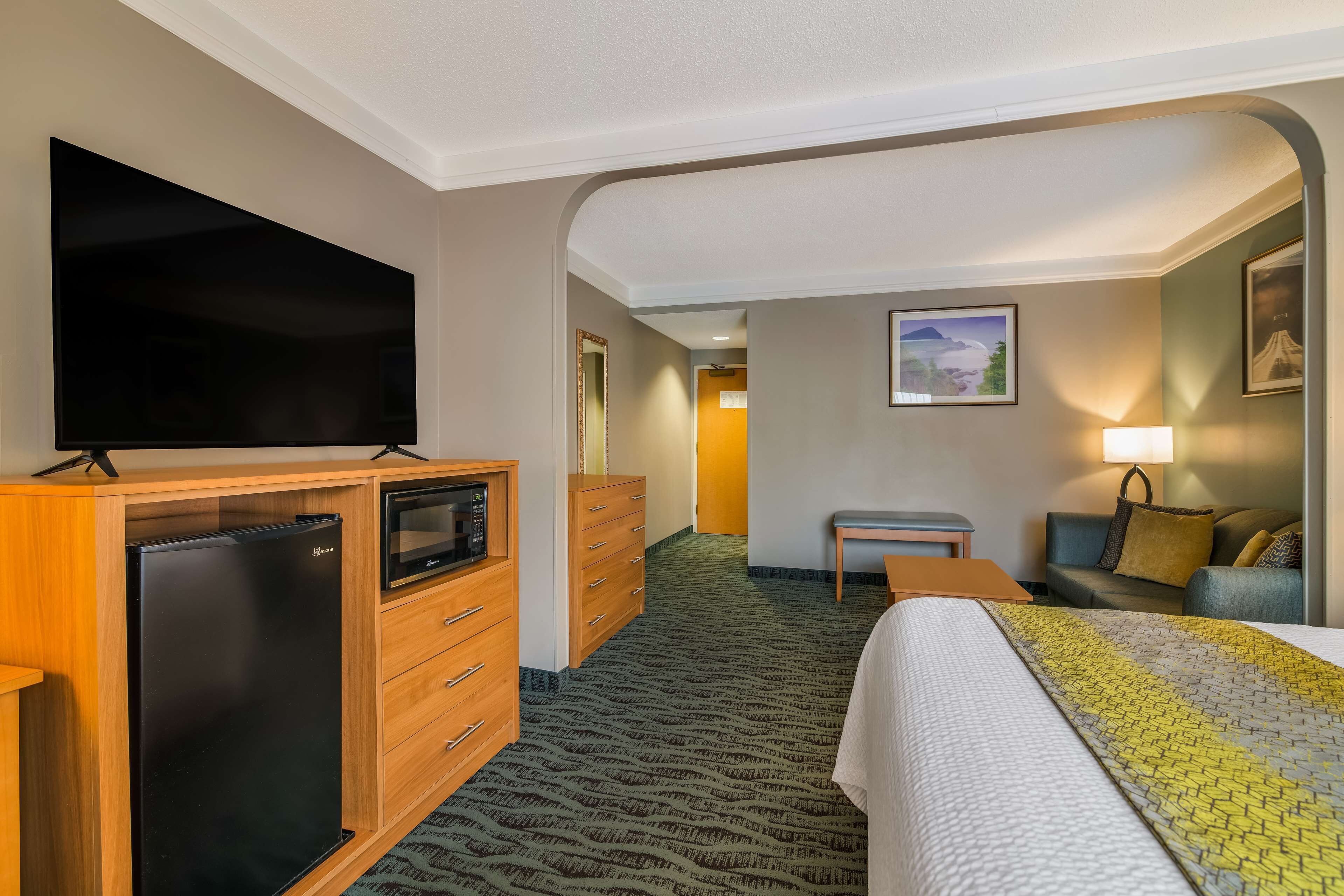 A hotel room with a flat-screen TV, a refrigerator, a wooden dresser, a bed with a patterned blanket, a small table, a sofa, and a lamp.