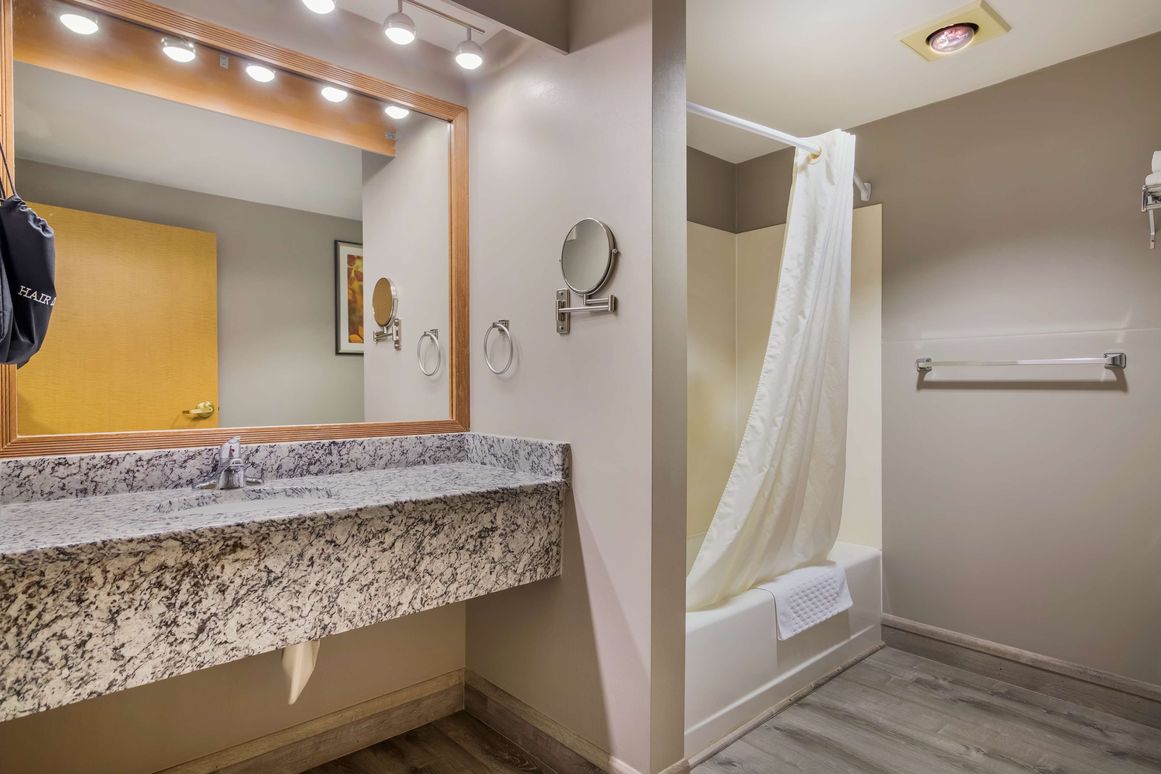 A bathroom with a granite countertop sink, a large mirror with lights, a towel rack, a toilet paper holder, and a shower with a white curtain.