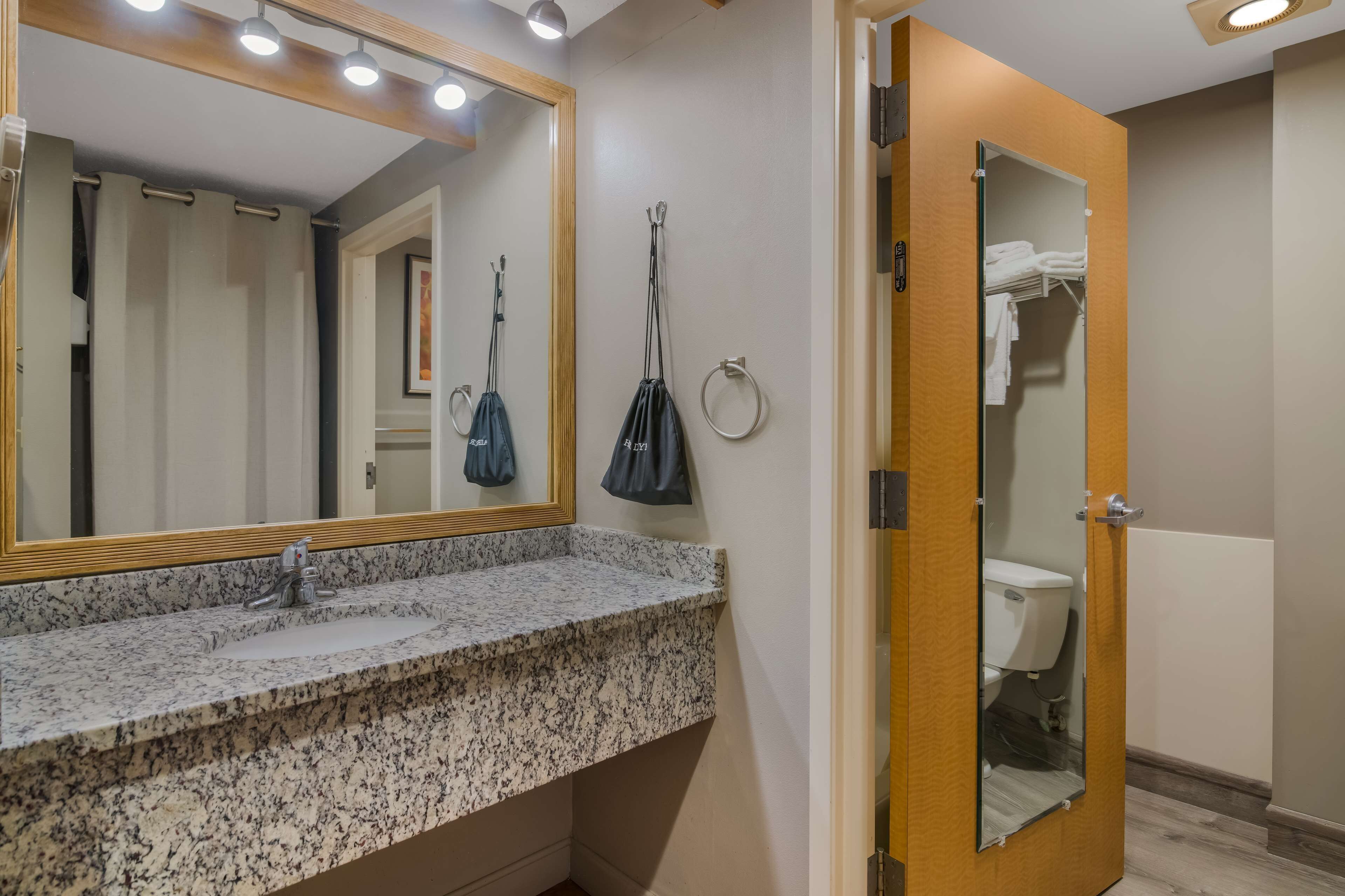 Une salle de bain avec un lavabo doté d’un plan de travail en granit, un miroir, un porte-serviettes et une porte en bois donnant accès aux toilettes.