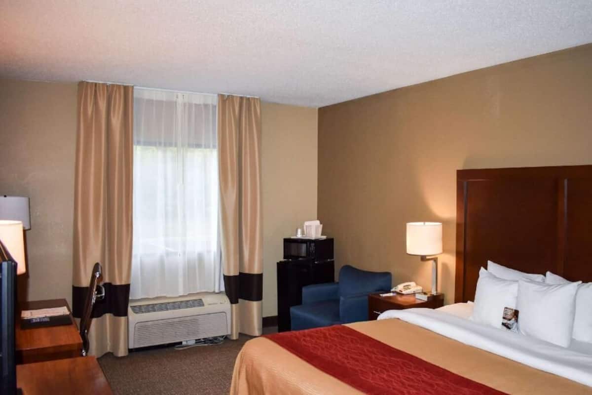 Ein Hotelzimmer mit Bett, Schreibtisch, Stuhl, Fernseher, Heizung, einem Fenster mit Vorhängen und einem Nachttisch mit Lampe.