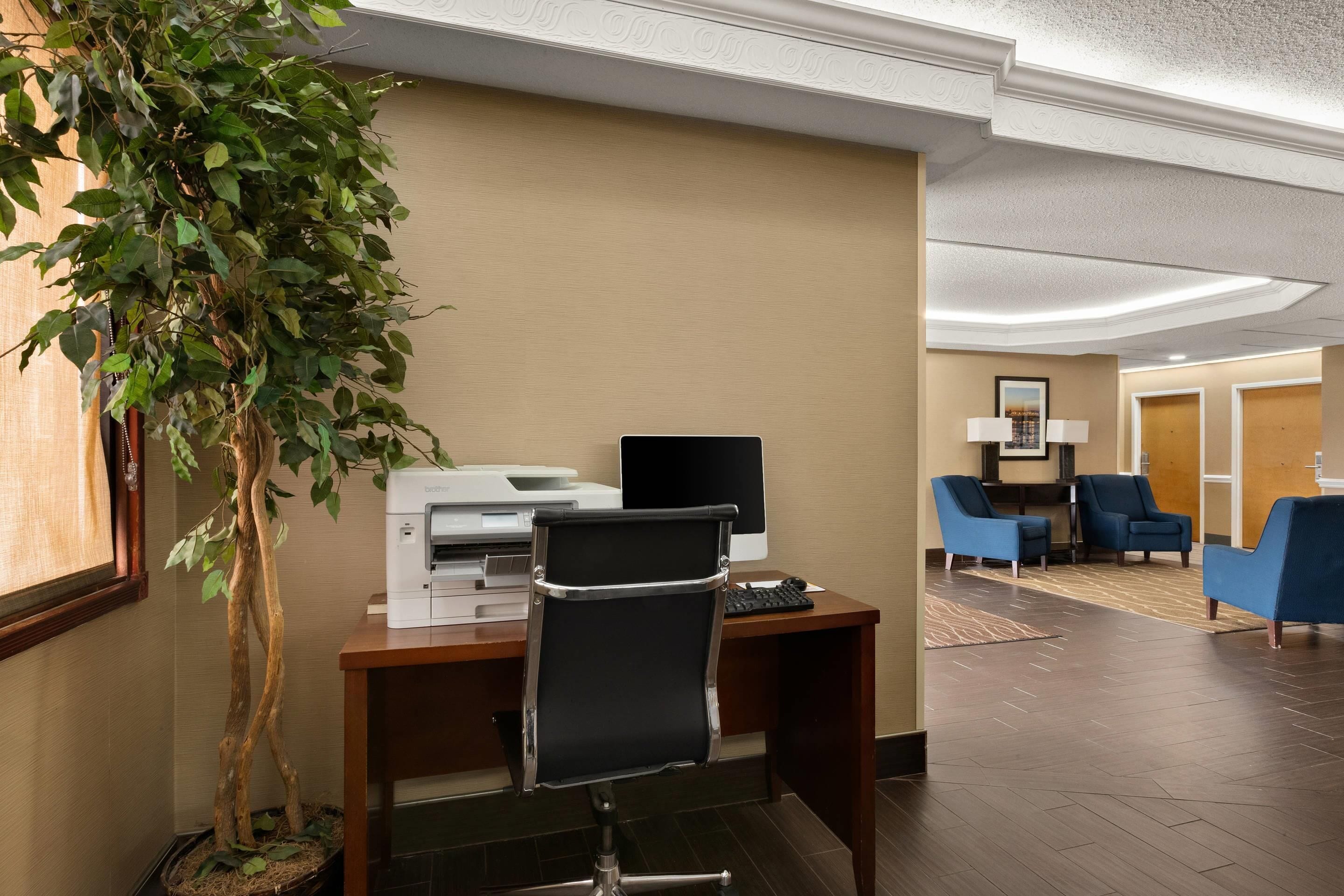 A hotel lobby with a reception desk, a computer, a printer, a potted plant, and blue chairs.