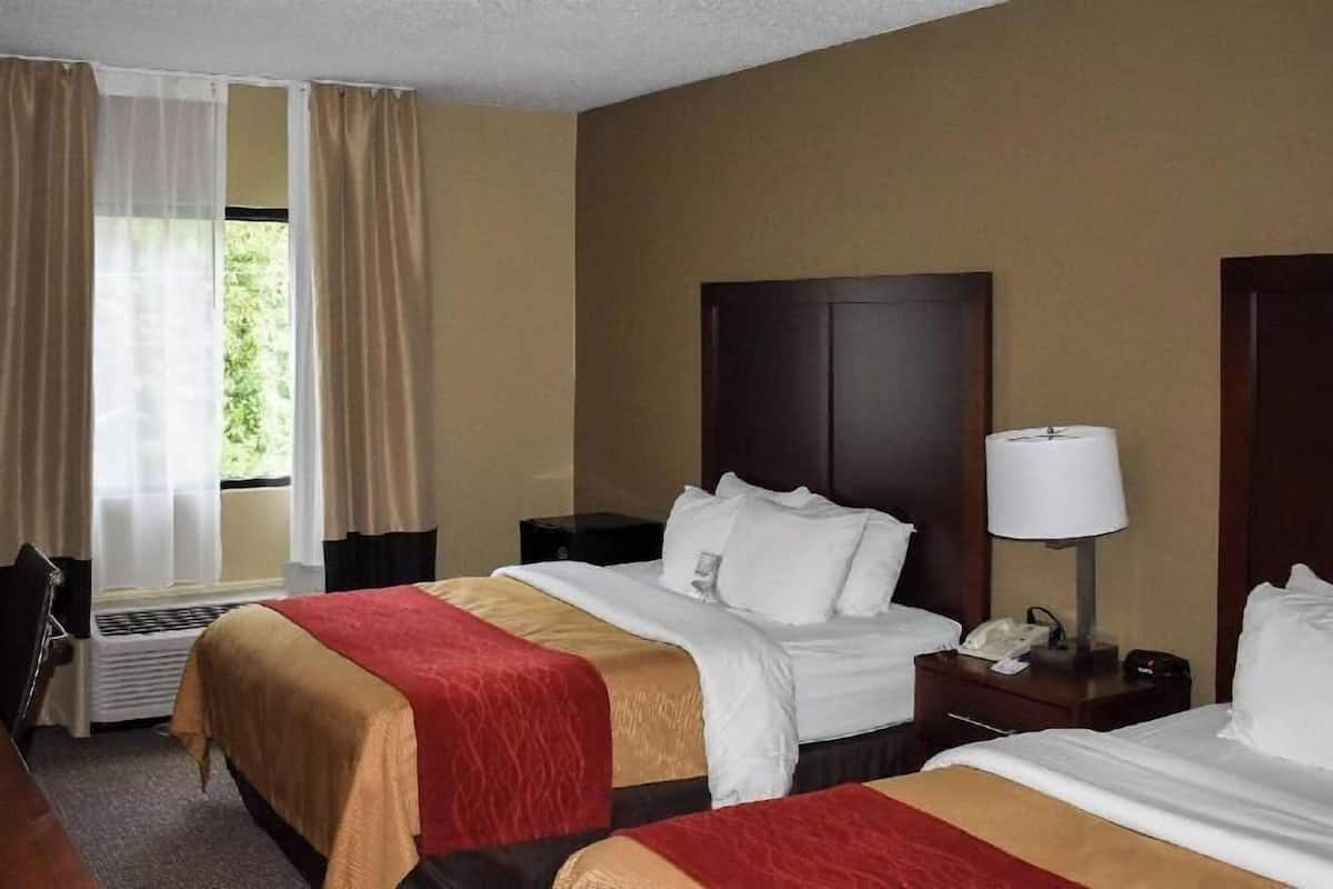 Ein Hotelzimmer mit zwei Betten, einem Schreibtisch, einem Stuhl, einem Fenster mit Vorhängen und einem Nachttisch mit Lampe.