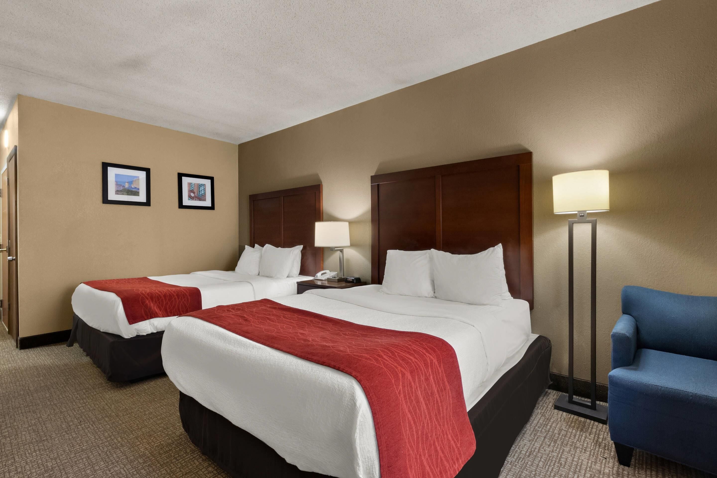 A hotel room with two beds, a desk, a chair, and a lamp.
