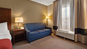 Una habitación de hotel con cama, sofá azul, mesita de noche con lámpara, ventana con cortinas y un calentador de pared.