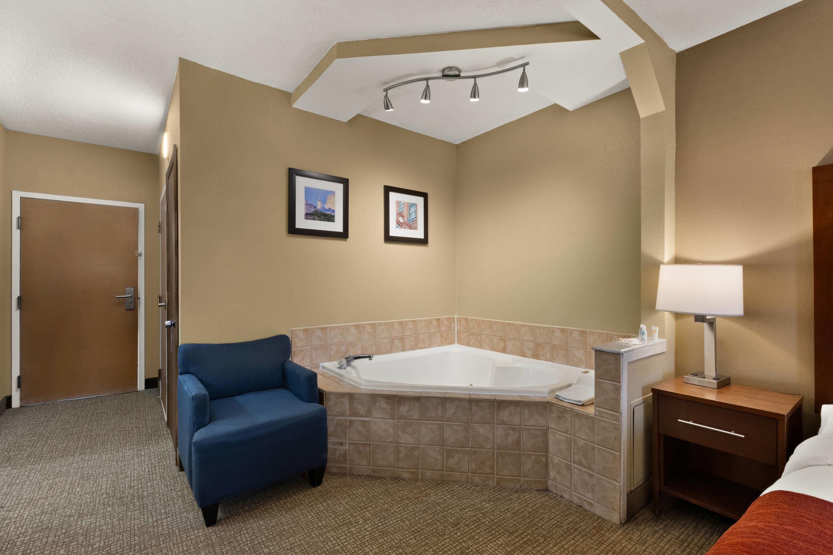 Habitación de hotel con una bañera grande, un sillón azul, una mesita de noche de madera con una lámpara y dos cuadros enmarcados en la pared.