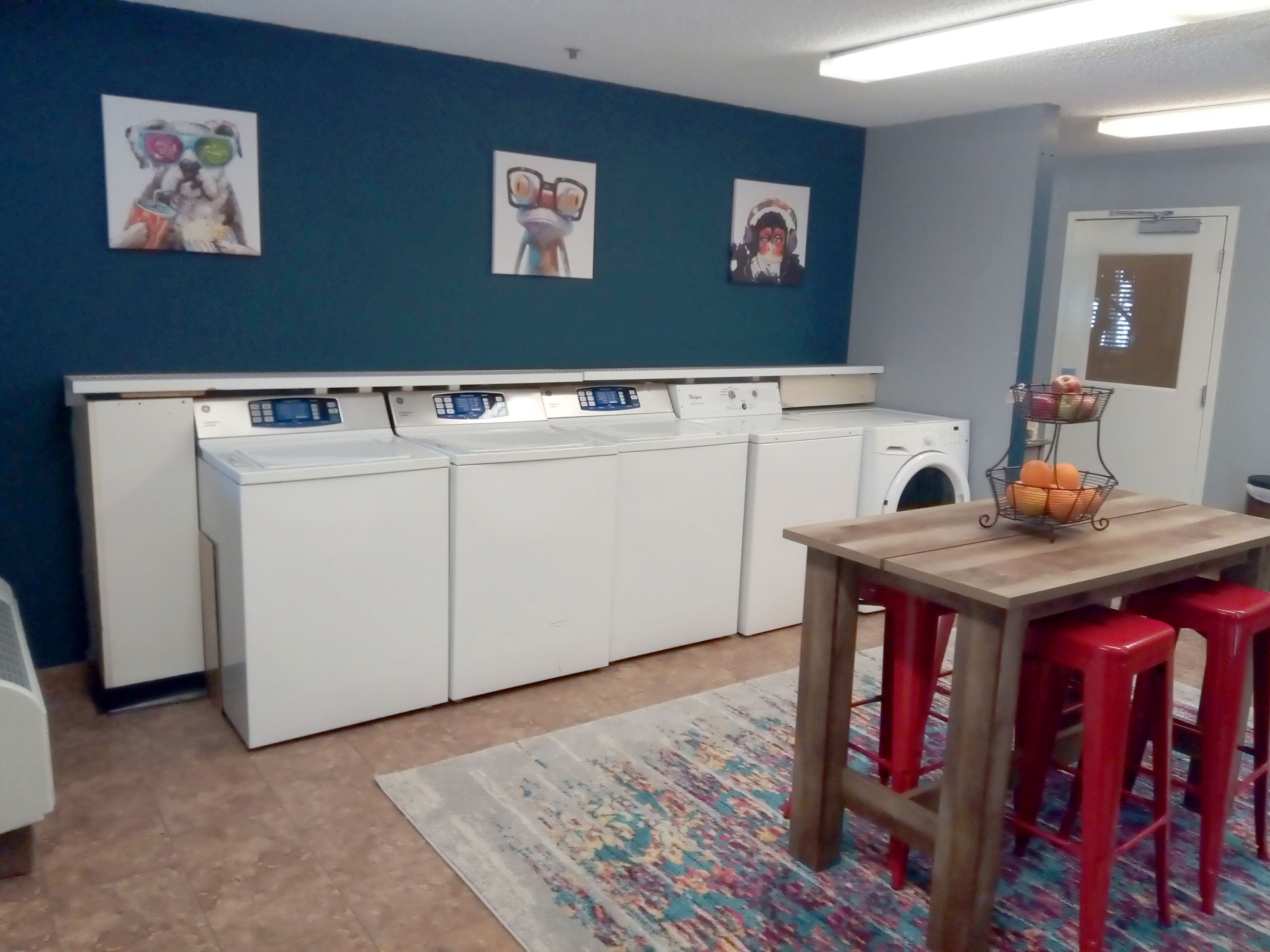 A laundry room with four white washing machines, a wooden table with two red stools, and a colorful rug on the floor.