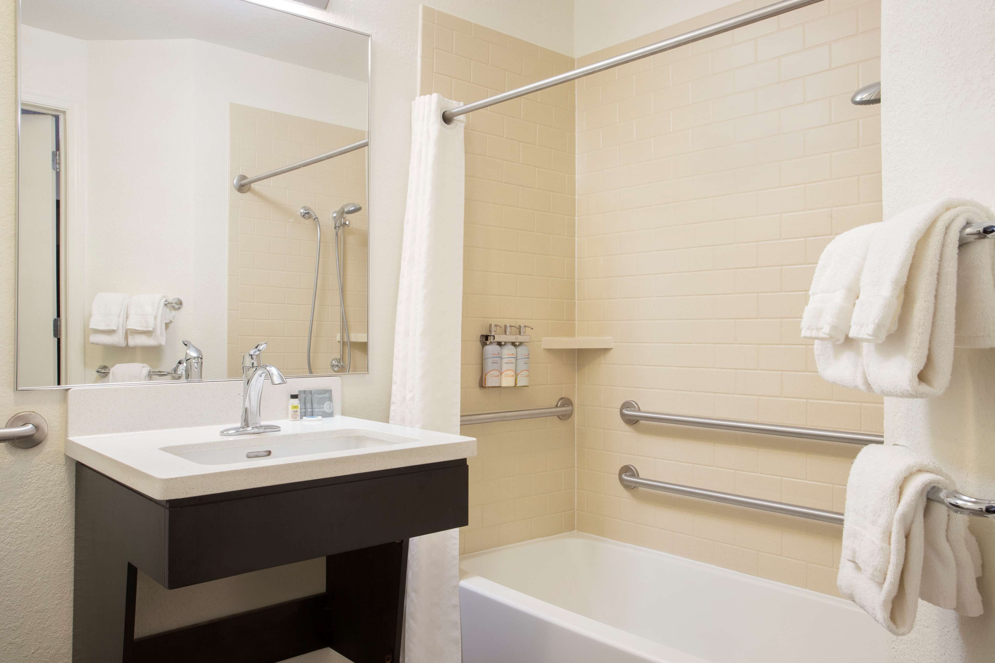 A bathroom with a sink, mirror, and bathtub. There are handrails and a shower curtain.