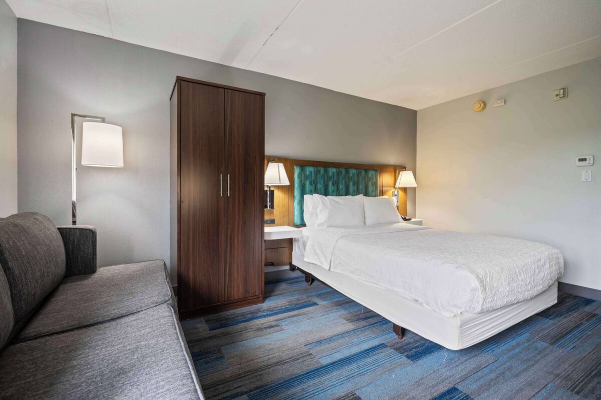 A hotel room with a bed, a wardrobe, a nightstand with lamps, and a sofa.