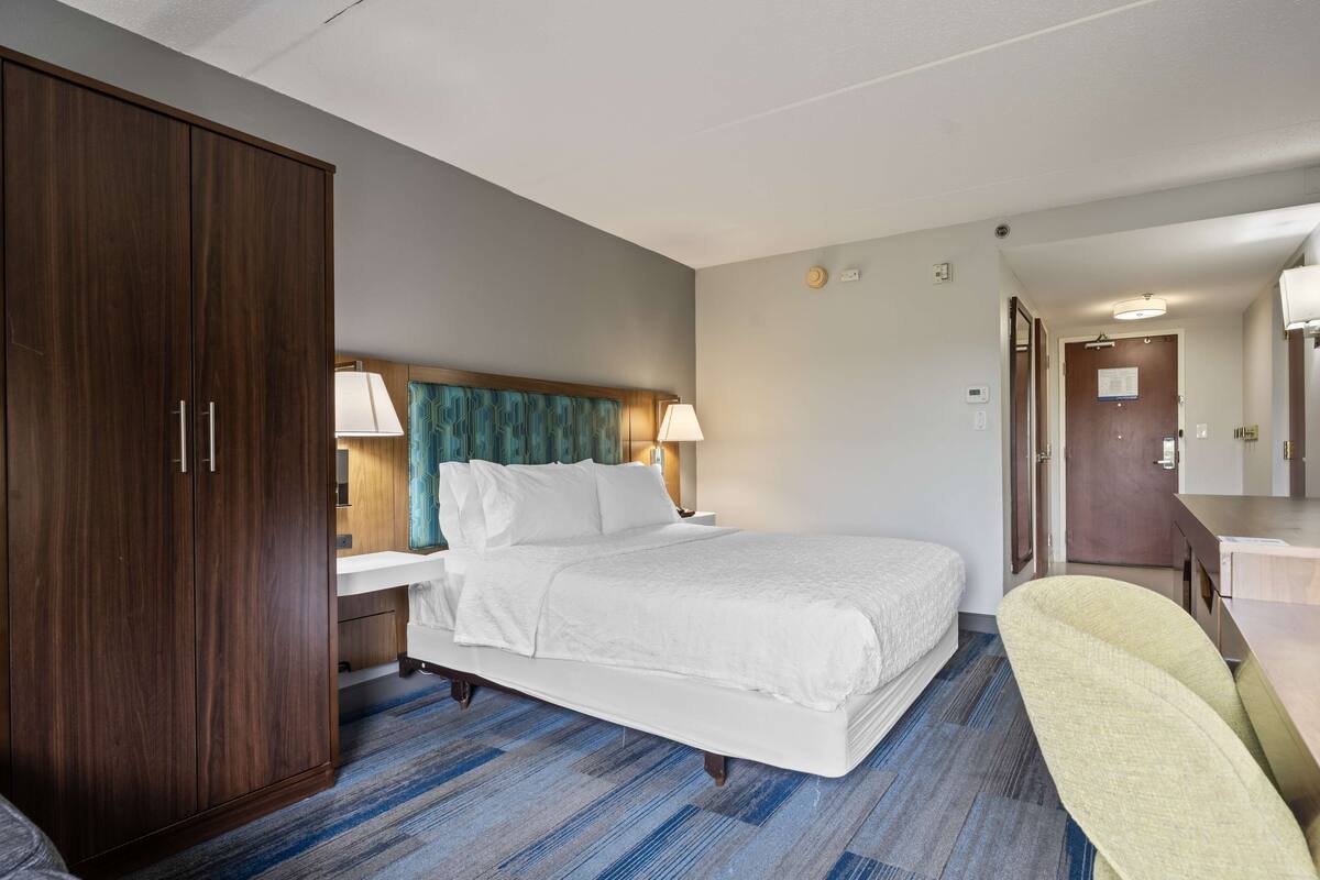 A hotel room with a large bed, a wooden wardrobe, a desk with a lamp, and a chair.