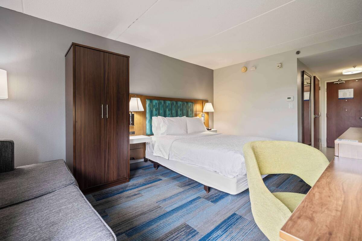 A hotel room with a bed, a wardrobe, a desk, a chair, and a lamp.