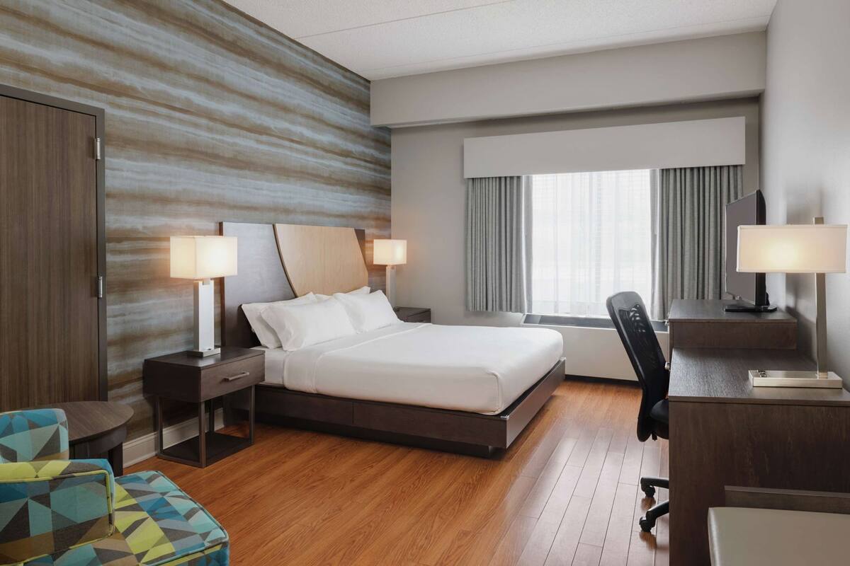 A hotel room with a bed, bedside tables, a desk, a chair, a window with curtains, and a wall with a textured pattern.