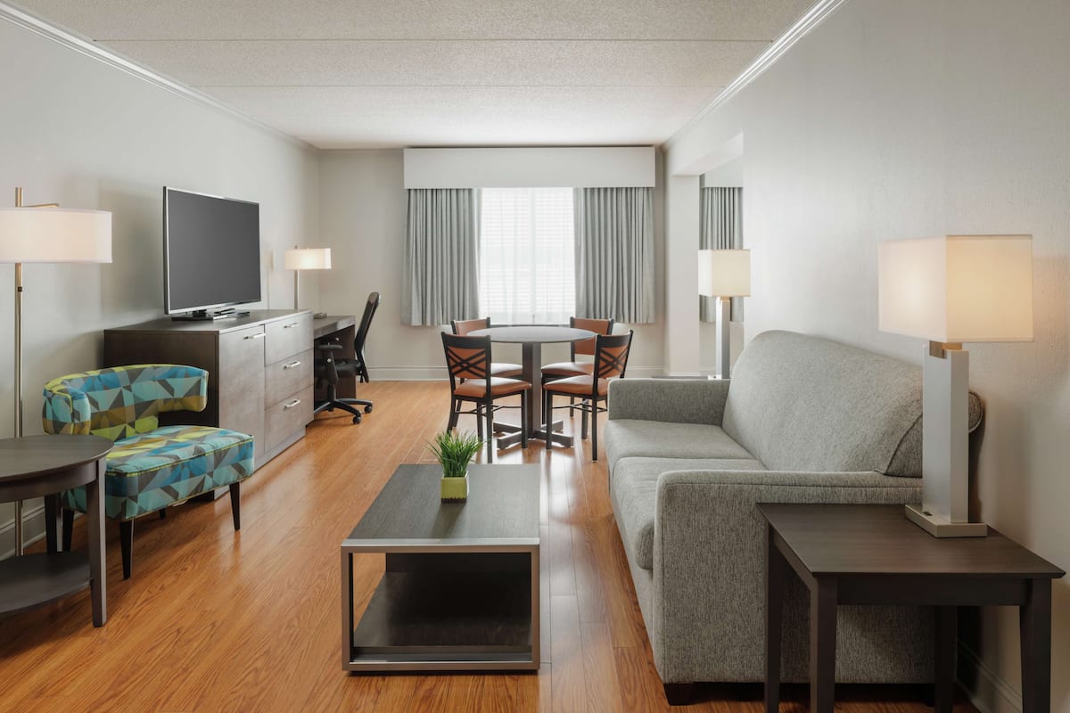 A modern hotel room with a flat-screen TV, a dining table with chairs, a sofa, a coffee table, and two lamps.