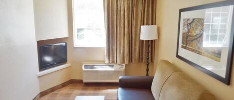 A hotel room with a brown leather sofa, a small wooden table, a flat-screen TV mounted on the wall, a standing lamp, a window with curtains, and a framed picture on the wall.