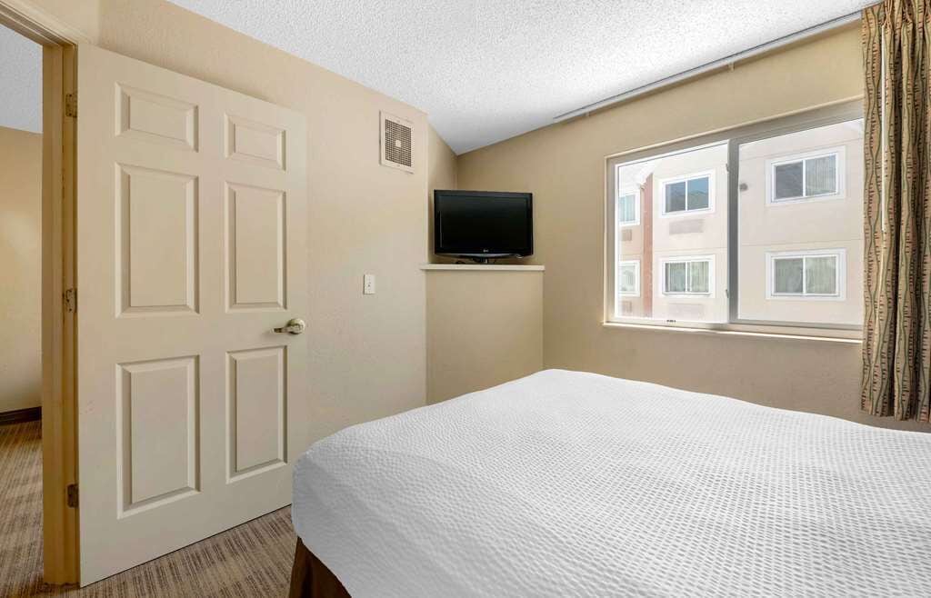 Habitació d'hotel amb un llit, una televisió sobre un suport, una finestra amb cortines i una porta.