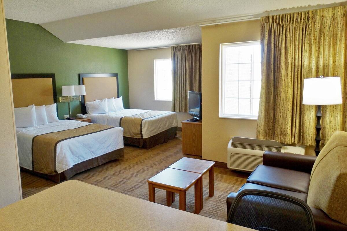 A hotel room with two beds, a desk, a chair, a TV, and an air conditioning unit.