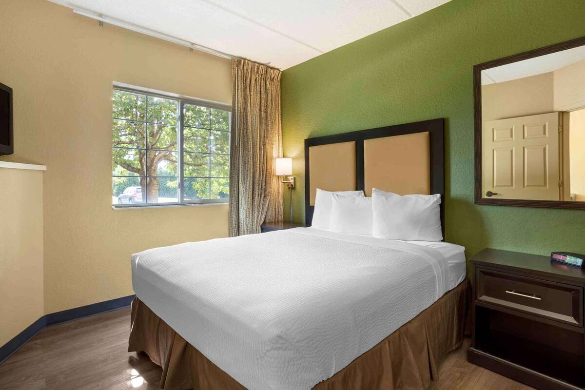 A hotel room with a bed, a nightstand, a lamp, a window with curtains, and a television on the wall.