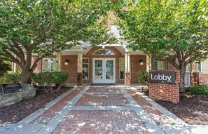A brick building entrance with a sign reading 'Lobby,' surrounded by trees and a paved walkway.